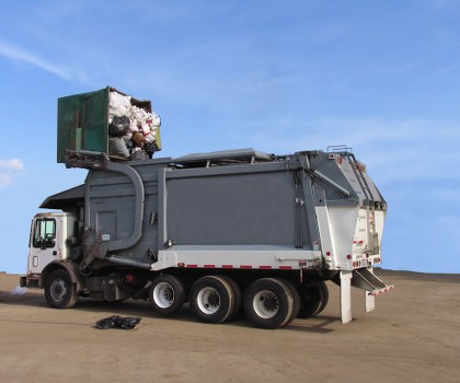 A large commercial rubbish collection truck parked on a paved surface under a clear blue sky. The truck has a white cab at the front and a substantial grey and silver waste containment body at the rear, designed for waste storage and compaction. The rear section features a hydraulic lifting mechanism with a partially opened green waste disposal bin positioned at the top, revealing cotton and paper waste inside. The vehicle is equipped with multiple axles, including six wheels visible along its length, and is oriented to the left side of the image. The environment appears to be an open area, possibly near an industrial or waste management site in Hampstead or surrounding London areas, and is clean with minimal foreground debris. The scene suggests active rubbish collection or waste disposal operations, consistent with commercial waste services provided by companies such as Commercial Waste Hampstead.