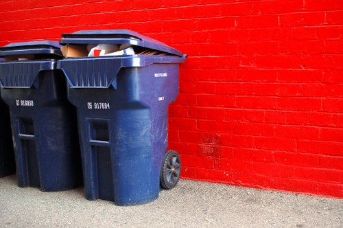 Two large dark blue plastic wheelie bins with closed lids are positioned on a paved surface in front of a bright red brick wall. The bins have textured surfaces and are fitted with black wheels at the back, with one bin slightly in front of the other. One of the bins has a partially visible white and red label on its side. The paved ground appears grey with a rough texture, and the background wall features uniformly laid red bricks with visible mortar lines. The overall scene suggests a typical waste collection area possibly associated with commercial or residential premises in Hampstead, where rubbish removal services such as those offered by Commercial Waste Hampstead might operate. The lighting is neutral and even, indicating natural daylight, and the setting appears clean and organized, suitable for business waste collection activities.