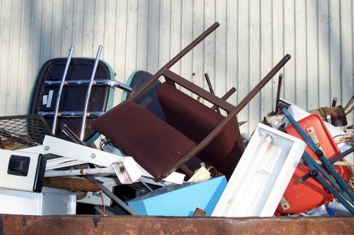 A pile of discarded household items and rubbish, including a maroon upholstered armchair with wooden legs, a white plastic storage container, and metal-framed furniture, all stacked on top of each other outdoors against a light-colored corrugated metal wall. The debris appears to be collected for rubbish removal services, with some items leaning and overlapping in a cluttered arrangement, situated on a paved surface. The scene is evenly lit, highlighting the different textures and colors of the various materials, with the focus on the waste items intended for disposal or clearance in a Hampstead residential or commercial area. The composition reflects typical rubbish collection scenarios managed by companies like Commercial Waste Hampstead.
