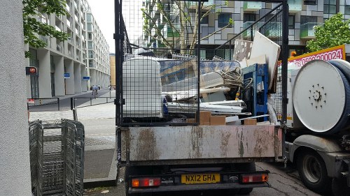 A small flatbed truck parked on a city street corner, loaded with various types of rubbish including wooden panels, metal scraps, and plastic containers. The truck's surface appears aged and slightly rusty, with a metal wire cage on one side for secure cargo. The rubbish extends to the back of the vehicle, spilling over the edges, and some items are partially leaning against the side. In the background, there are modern residential buildings with multiple windows and balconies, along with a section of pavement and a pedestrian crossing visible at the intersection. The scene is captured during daylight hours, with natural lighting highlighting the textures of the debris and the urban environment, reflecting typical rubbish removal services offered by companies like Commercial Waste Hampstead in the Hampstead area.