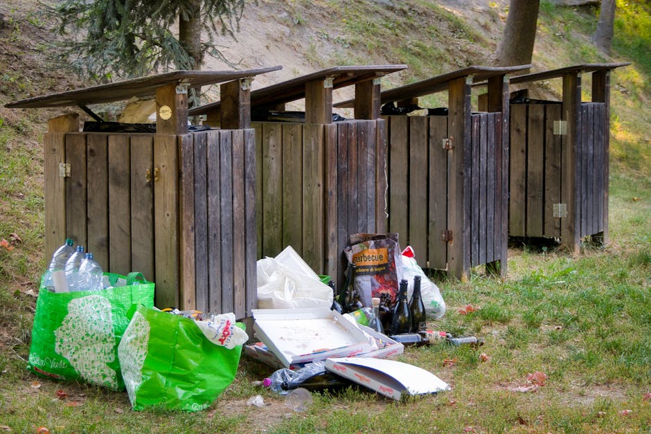 The image shows four wooden outdoor trash bins with sloped roofs, positioned side by side on a grassy area with a slight incline. The bins are weathered, dark brown, with visible wood grain and metal 